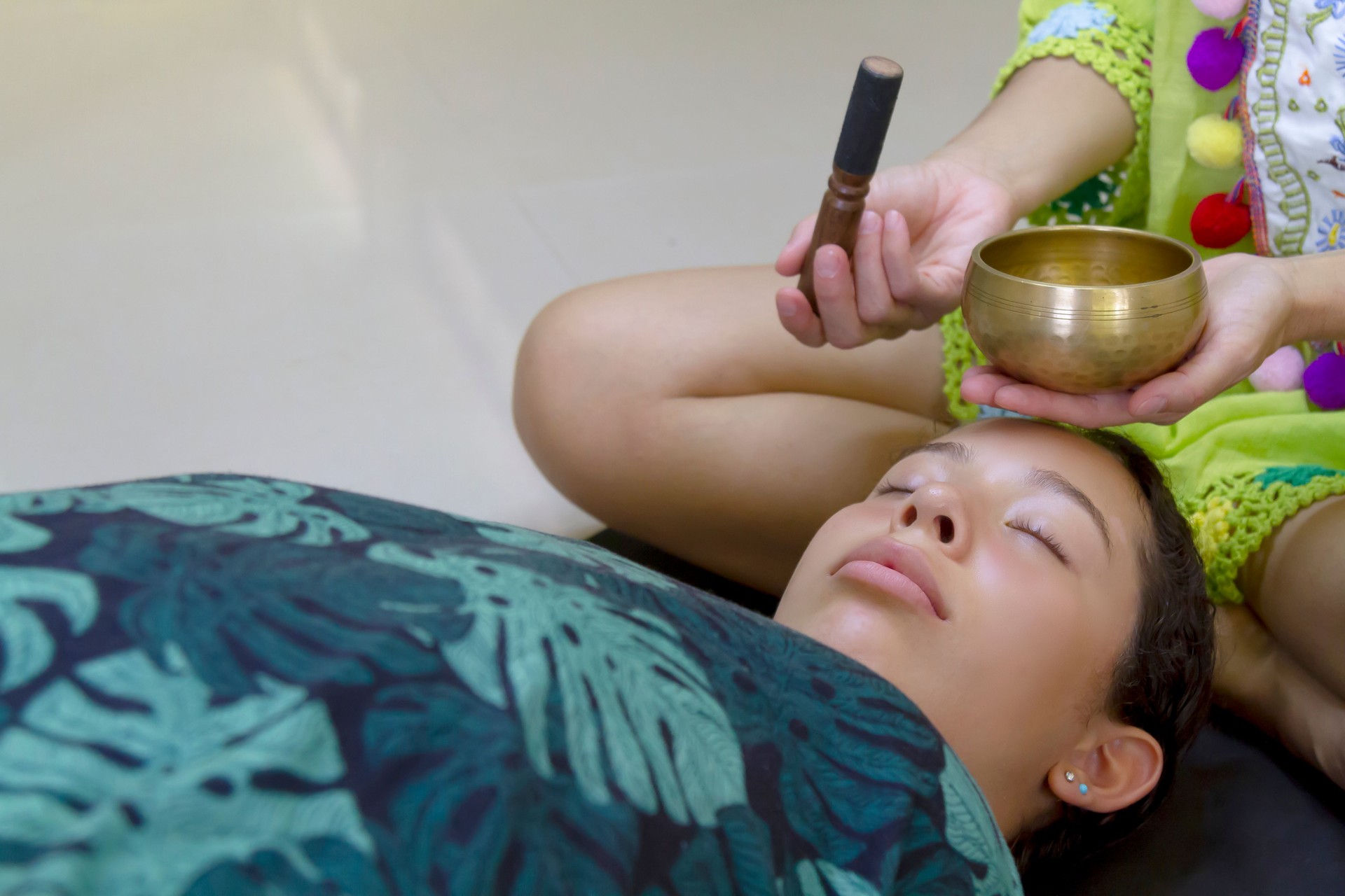 A single athletic middle age young active latin Spiritual Seeker woman practicing yoga and Meditating while playing on Tibetan singing bowl as a therapy to another young teenager woman inside a studio.